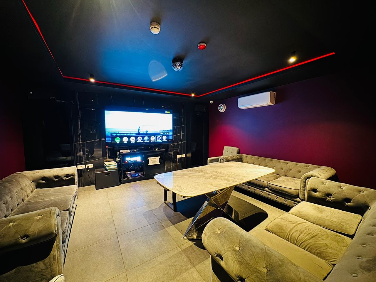 Inside a HANJAN karaoke room: sofa seating around a low table, a large wall screen and a mic console, red light along the ceiling